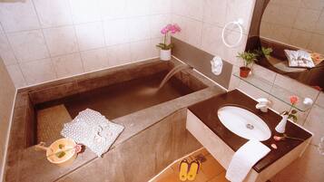 Un baño con bañera, lavamanos, espejo, toalla, planta en maceta y un par de pantuflas.