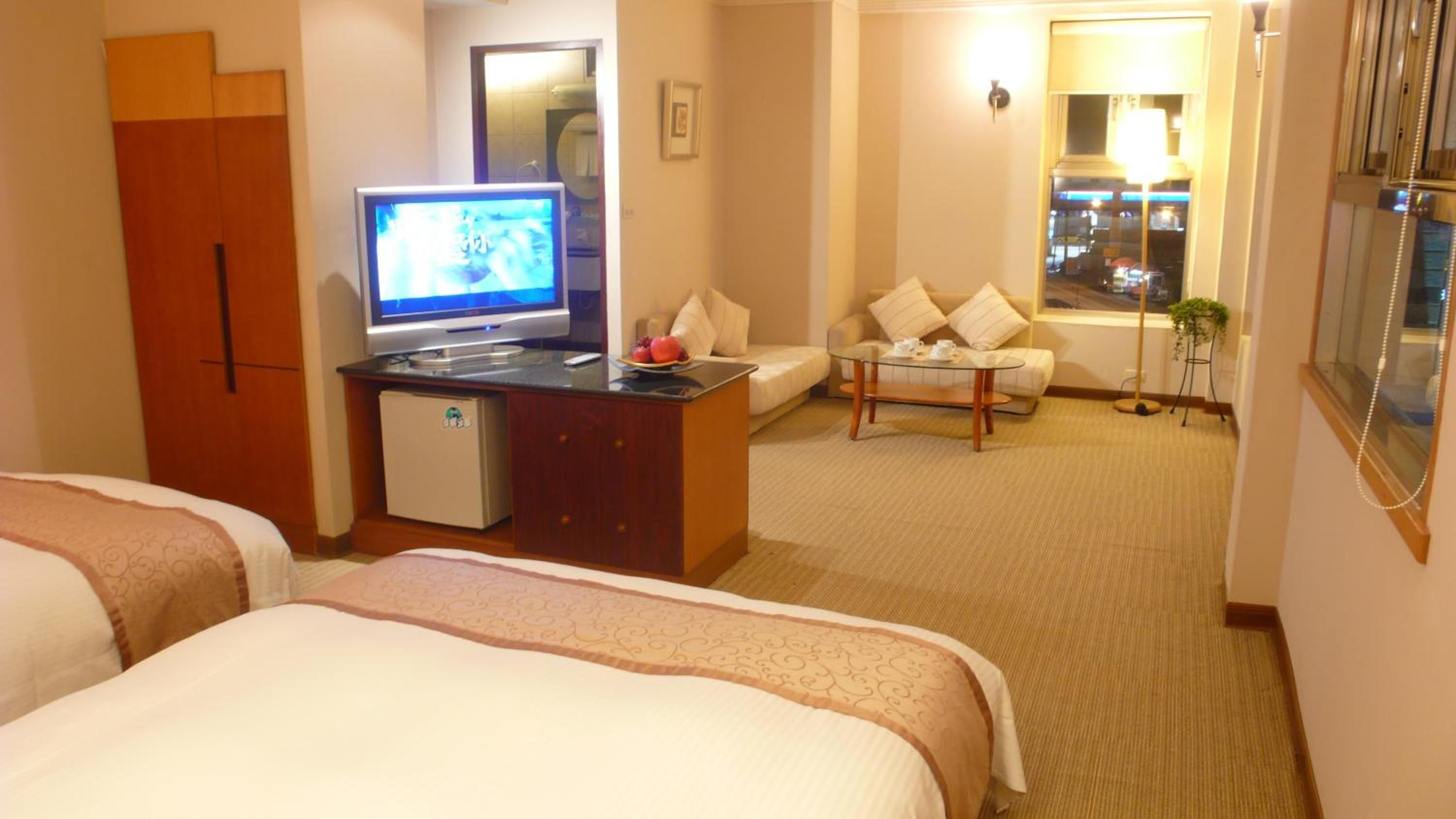 Habitación de hotel con dos camas, un escritorio con televisor, un refrigerador pequeño, un sofá y una ventana con vistas al exterior.