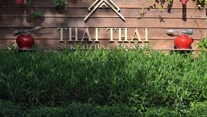 Une enseigne en bois portant le nom « THAI THAI » et, en dessous, les mots « SUKKHOHIAL RESORT ».