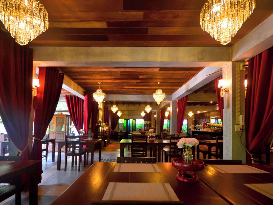 Un restaurante con vigas de madera en el techo, cortinas rojas y candelabros. Las mesas están dispuestas con posavasos y centros de mesa.