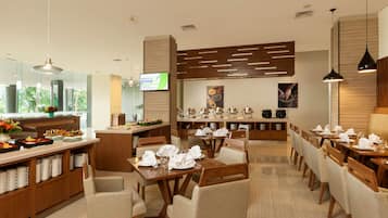 A hotel dining area with multiple tables set for guests, a buffet counter with food items, and a television screen displaying a welcome message.