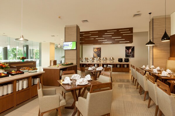 Un comedor de hotel con varias mesas dispuestas para los huéspedes, una barra de buffet con alimentos y una pantalla de televisión mostrando un mensaje de bienvenida.
