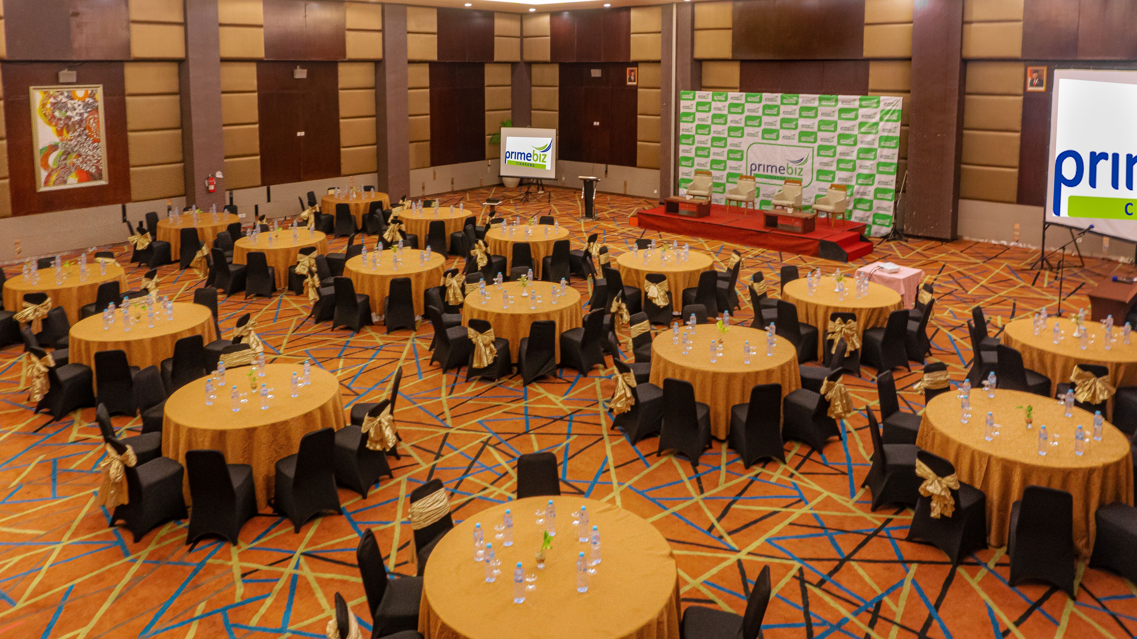 A conference room with multiple round tables, each with chairs and water bottles. A presentation screen is visible in the background.