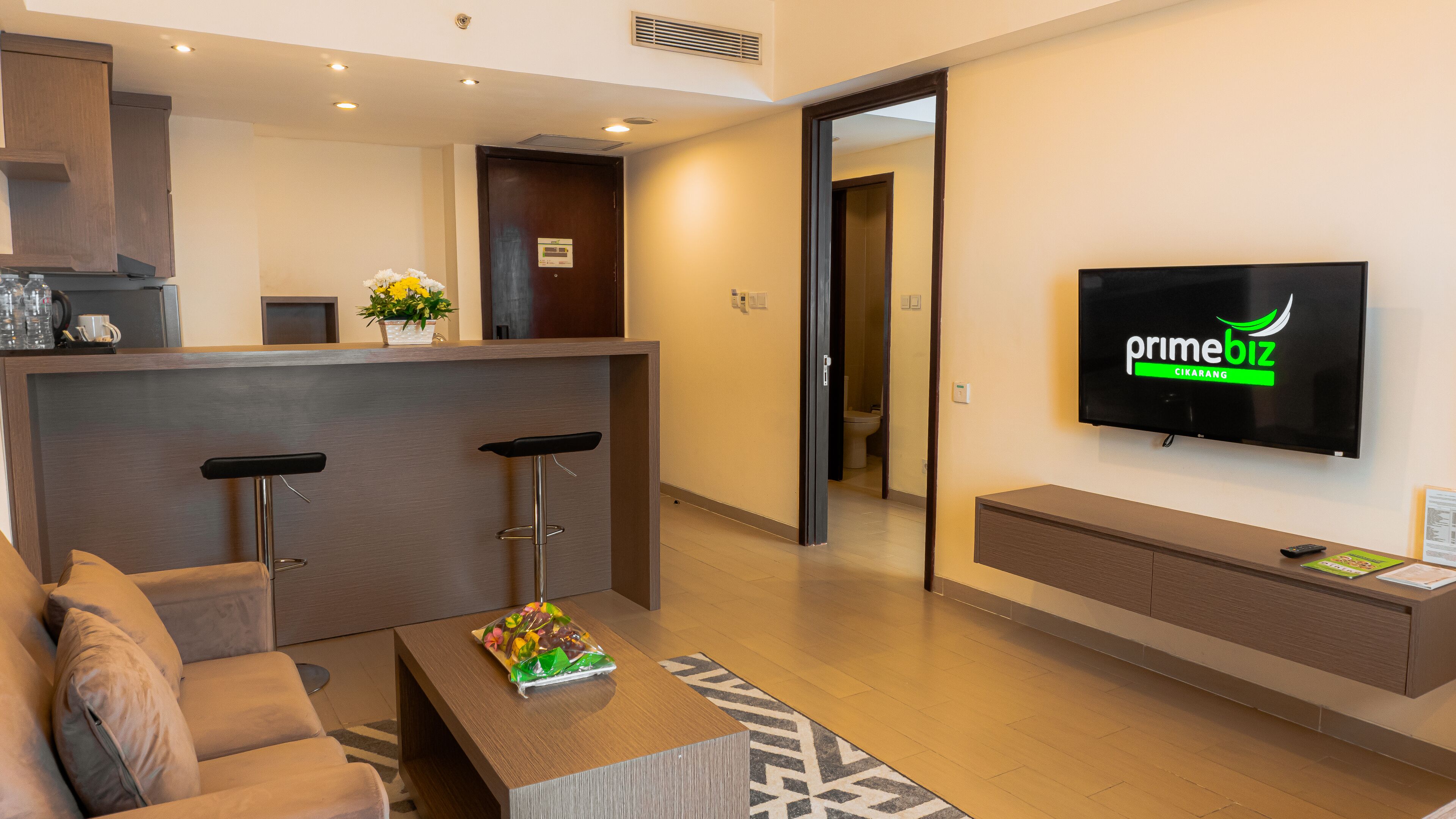 Un vestíbulo de hotel moderno con mostrador de recepción, dos taburetes de bar, un sofá, una mesa de centro con un objeto decorativo, un televisor de pantalla plana y un armario de madera.