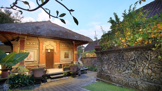 A traditional wooden house with a thatched roof, a stone wall with carvings, and a paved pathway leading to the entrance. - Arnawa Bungalow (Ubud)