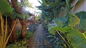 A narrow pathway flanked by large tropical plants and trees, leading towards a building with a tiled roof. - Arnawa Bungalow (Ubud)