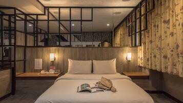 A hotel room with a wooden bed, two bedside lamps, a nightstand with a phone and a magazine, and a view of the interior through a large window.