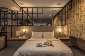 A hotel room with a wooden bed, two bedside lamps, a nightstand with a phone and a magazine, and a view of the interior through a large window.
