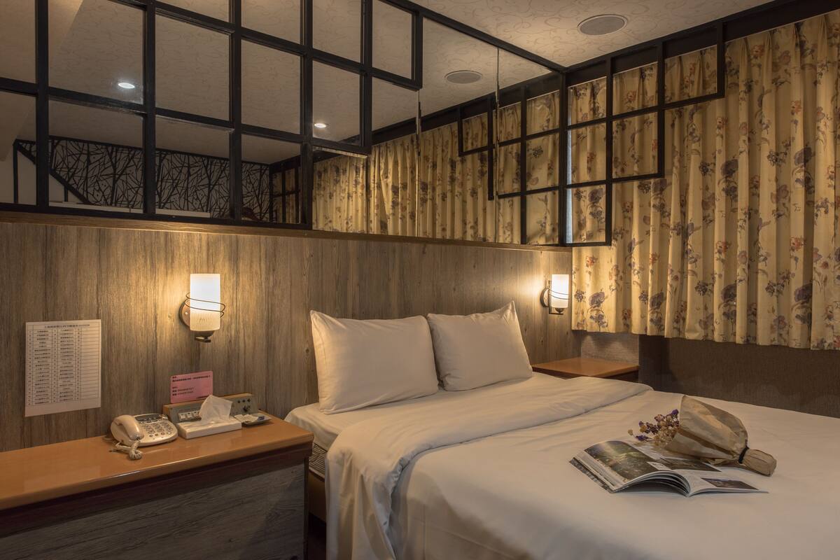 A hotel room with a wooden headboard, a bed with white linens, a bedside table with a telephone and a sign, and a wall-mounted light fixture.