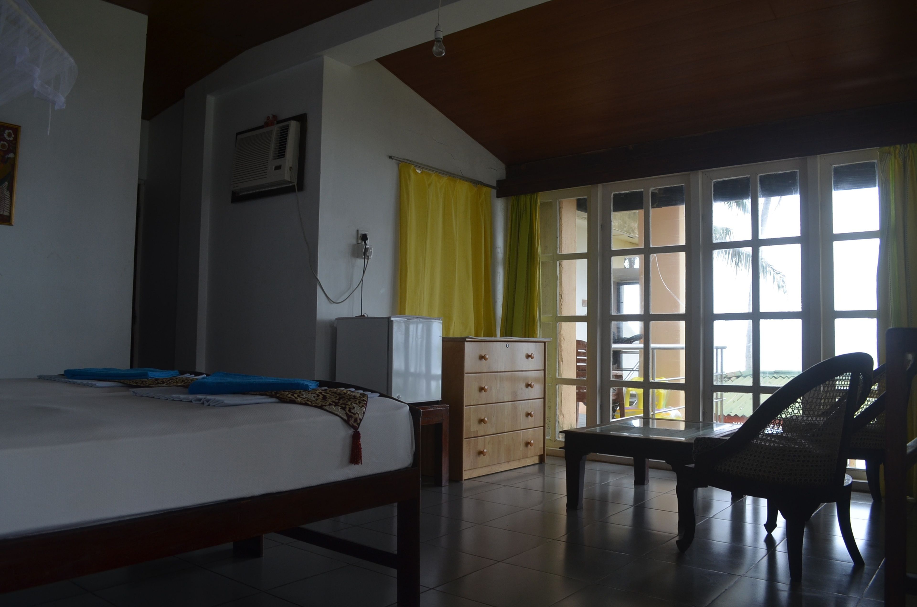 A room with a bed, a wooden dresser, a desk, and a chair. There is a window with yellow curtains and a view of the outside.