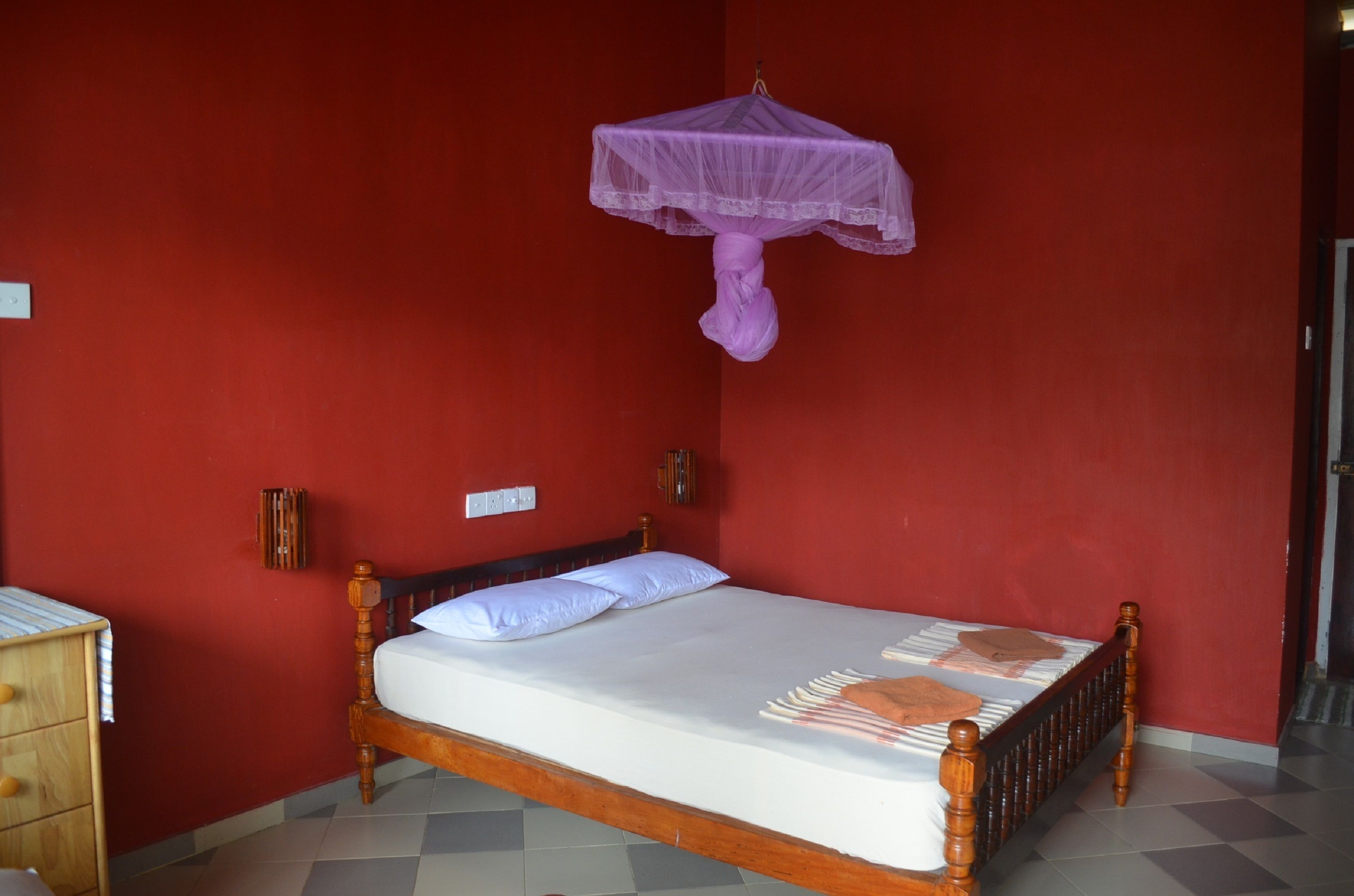 Une chambre avec un lit simple, un cadre en bois, un matelas blanc et une moustiquaire violette.