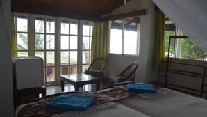 Una habitación con cama, una mesa y una silla. Hay ventanas con cortinas y vistas a palmeras afuera.
