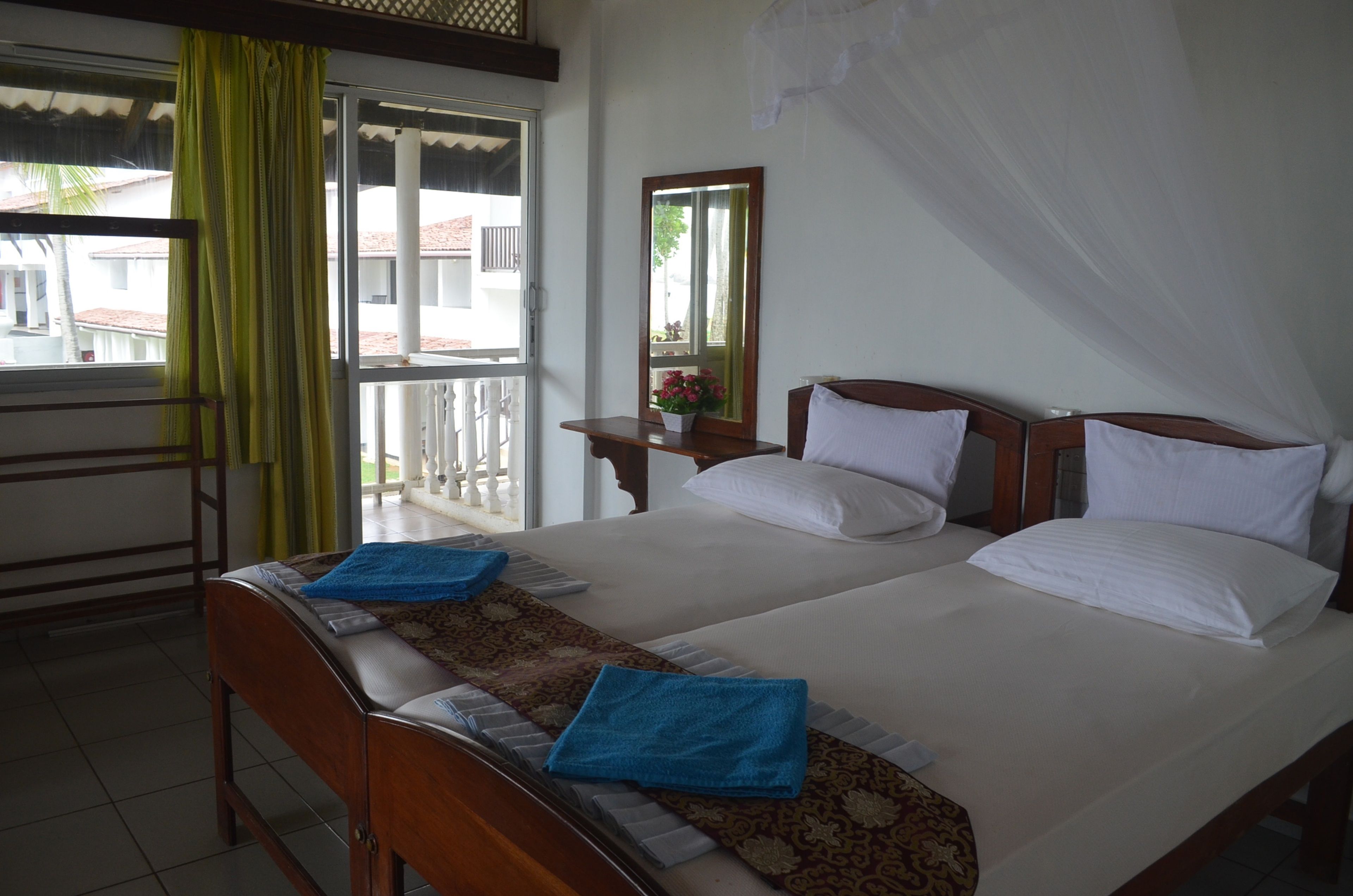 A hotel room with two beds, a window with curtains, a bedside table with a mirror, and a view of the outside.