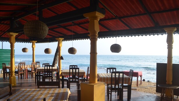 Un área de comedor al aire libre con mesas y sillas, techo rojo y macetas colgantes.