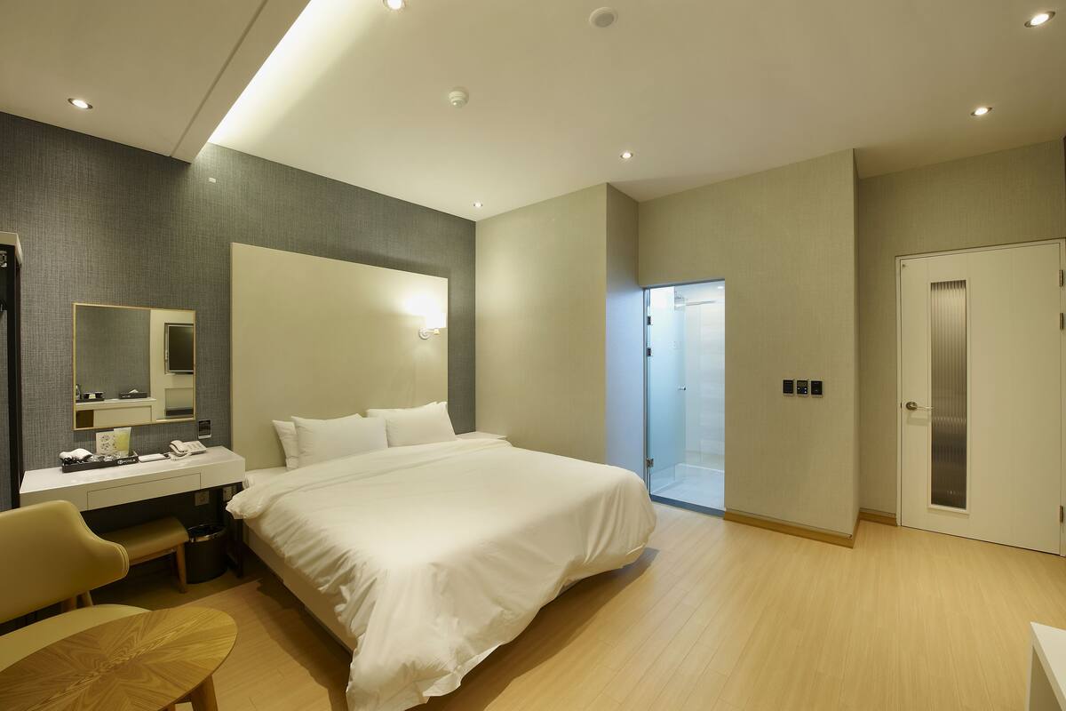 A modern hotel room with a large bed, a desk with a chair, a small table, and a mirror.