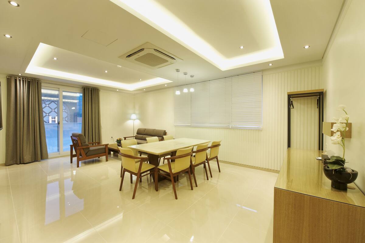 A modern hotel room with a large dining table, comfortable seating, a large window with curtains, and a reception desk with a vase of flowers.