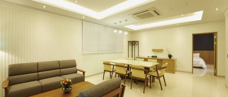A modern living room with a sofa, dining table, and chairs. There is a small table with a vase of flowers. The room has a large window with blinds and a ceiling with recessed lighting.