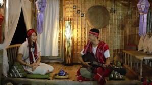 A traditional wooden hut interior with a thatched roof, a woven basket hanging on the wall, and a person playing a stringed instrument.