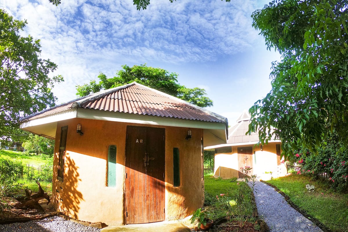 A small, single-story building with a tiled roof and a wooden door, surrounded by lush greenery and a gravel path.