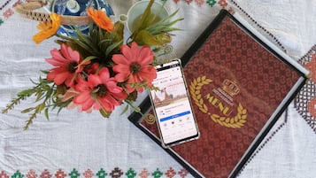 A table with a decorative tablecloth, a vase with flowers, a smartphone displaying a restaurant menu, and a menu book.