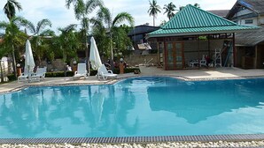 A swimming pool with lounge chairs and umbrellas, surrounded by palm trees and a building with a green roof.