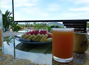 A table with a fruit platter, a glass of orange juice, and a mug on a balcony with a view of the sky and greenery. - Baan Georges Hotel (Sukhothai)