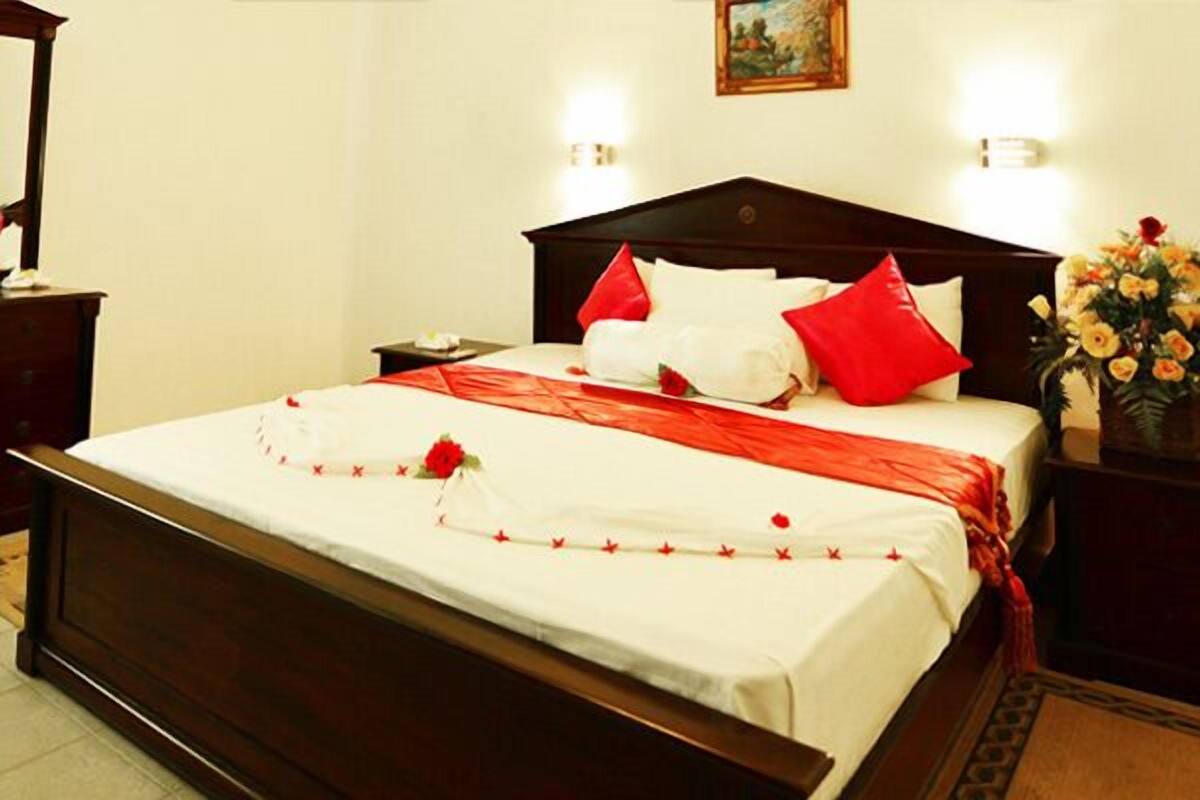 Uma cama bem arrumada com lençóis brancos, almofadas vermelhas e um objeto decorativo vermelho. Há uma mesa de cabeceira com um abajur e um vaso de flores.