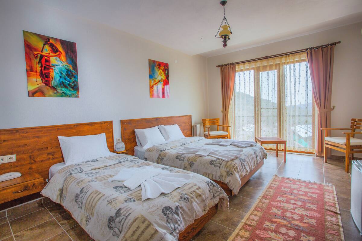 Une chambre d’hôtel avec deux lits, une tête de lit en bois, une table de chevet, une chaise, une table et une grande fenêtre avec des rideaux.