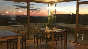 Ein Essbereich mit Tisch für vier Personen, große Fenster mit Blick auf den Sonnenuntergang und ein Blumentopf am Fensterbrett.