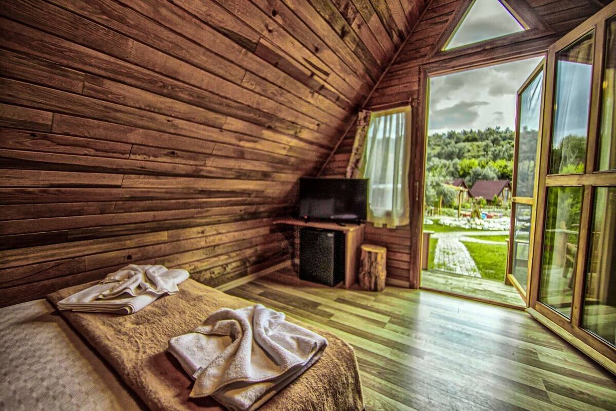 Une chambre dans un chalet en bois, équipée d’un lit, d’une télévision et d’une grande fenêtre donnant sur un jardin et des bâtiments.