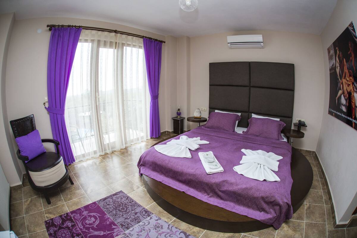 Une chambre à coucher moderne avec un grand lit, une literie violette, un fauteuil et une vue sur l’extérieur.
