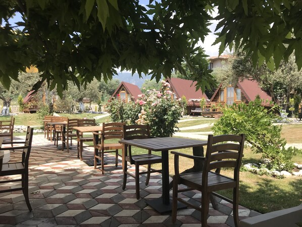 Un espace patio avec des tables et des chaises en bois, entouré de verdure et offrant une vue sur des cabanes rustiques.