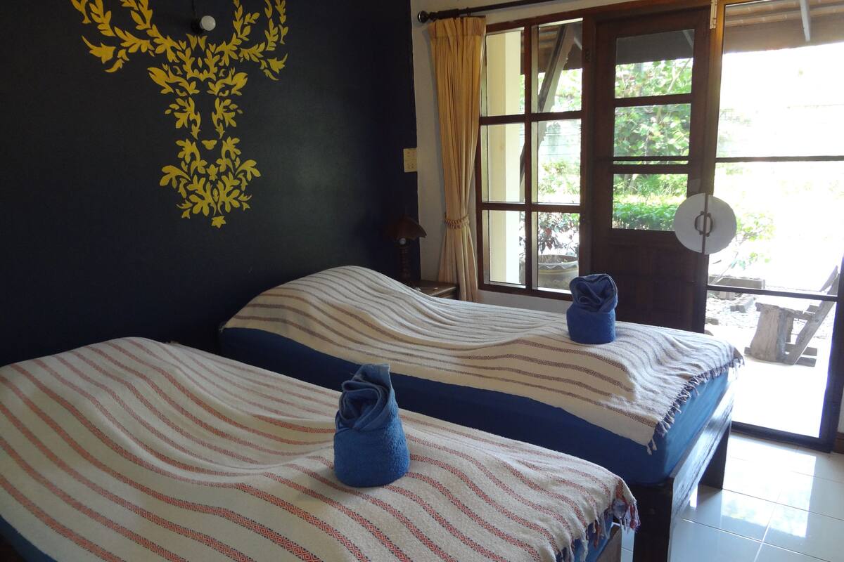 A room with two beds, each with a striped blanket and a blue towel. There is a window with curtains, a wall decoration, and a door leading outside.