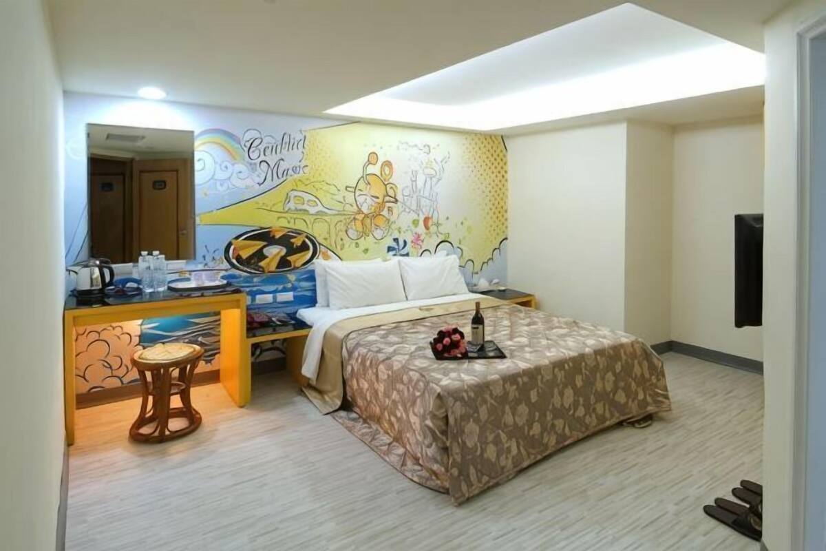 Une chambre d’hôtel comprenant un lit, un bureau avec un ordinateur, une chaise, un petit tabouret, une décoration murale et une porte.