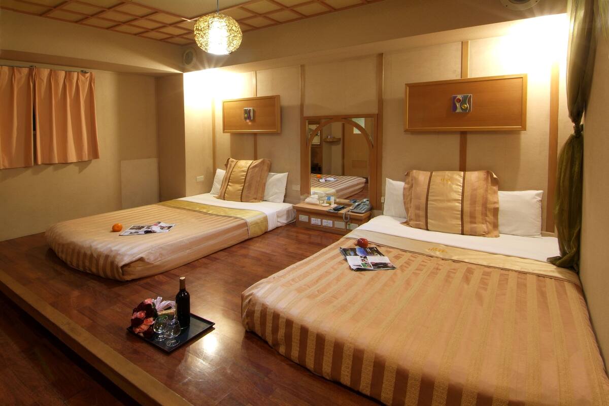 Une chambre d’hôtel avec deux lits simples, un plancher en bois, une petite table avec une bouteille et des verres, et un miroir au mur.
