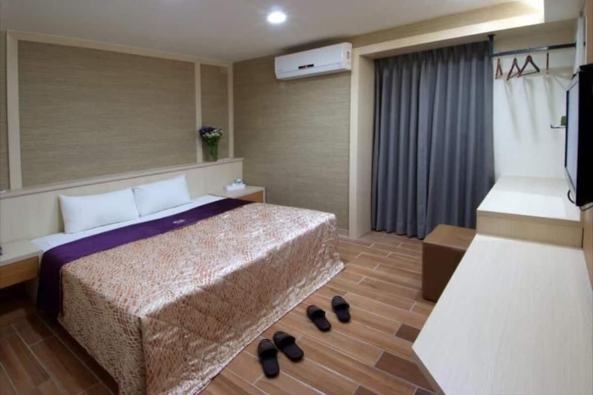 Une chambre d’hôtel comprenant un lit, une table de chevet, une télévision, un climatiseur, une fenêtre avec des rideaux et un porte-chaussures.