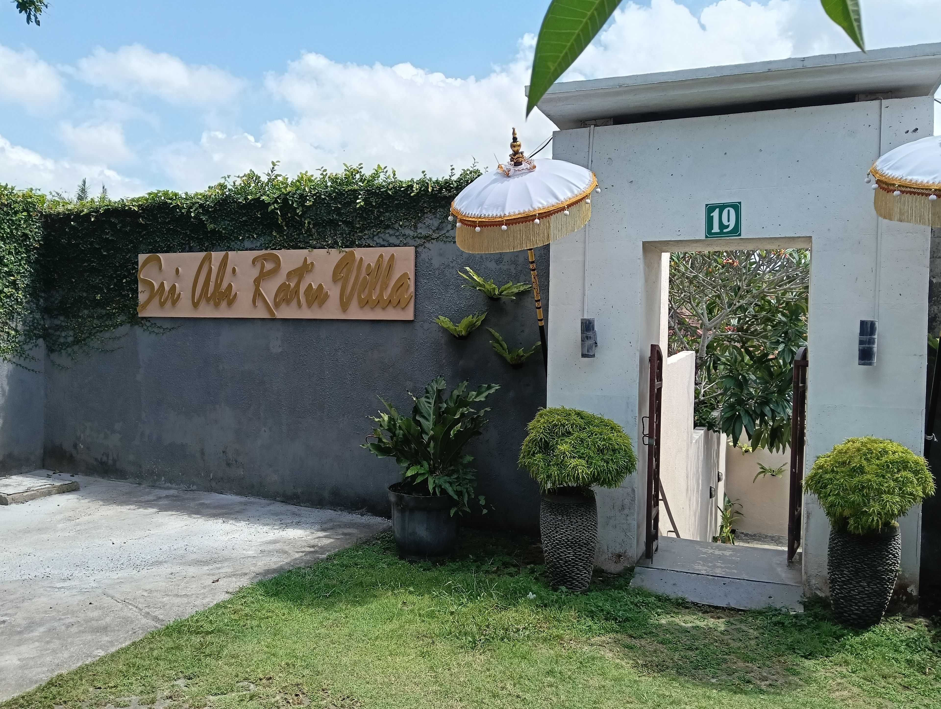 A villa entrance with a sign reading 'Sri Abhi Ratu Villa,' a concrete wall with greenery, and a number '19' on the door.
