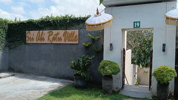 A villa entrance with a sign reading 'Sri Abhi Ratu Villa,' a concrete wall with greenery, and a number '19' on the door.