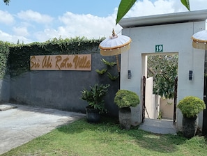 Entrada a una villa con un letrero que dice 'Sri Abhi Ratu Villa', una pared de concreto con follaje y el número '19' en la puerta.