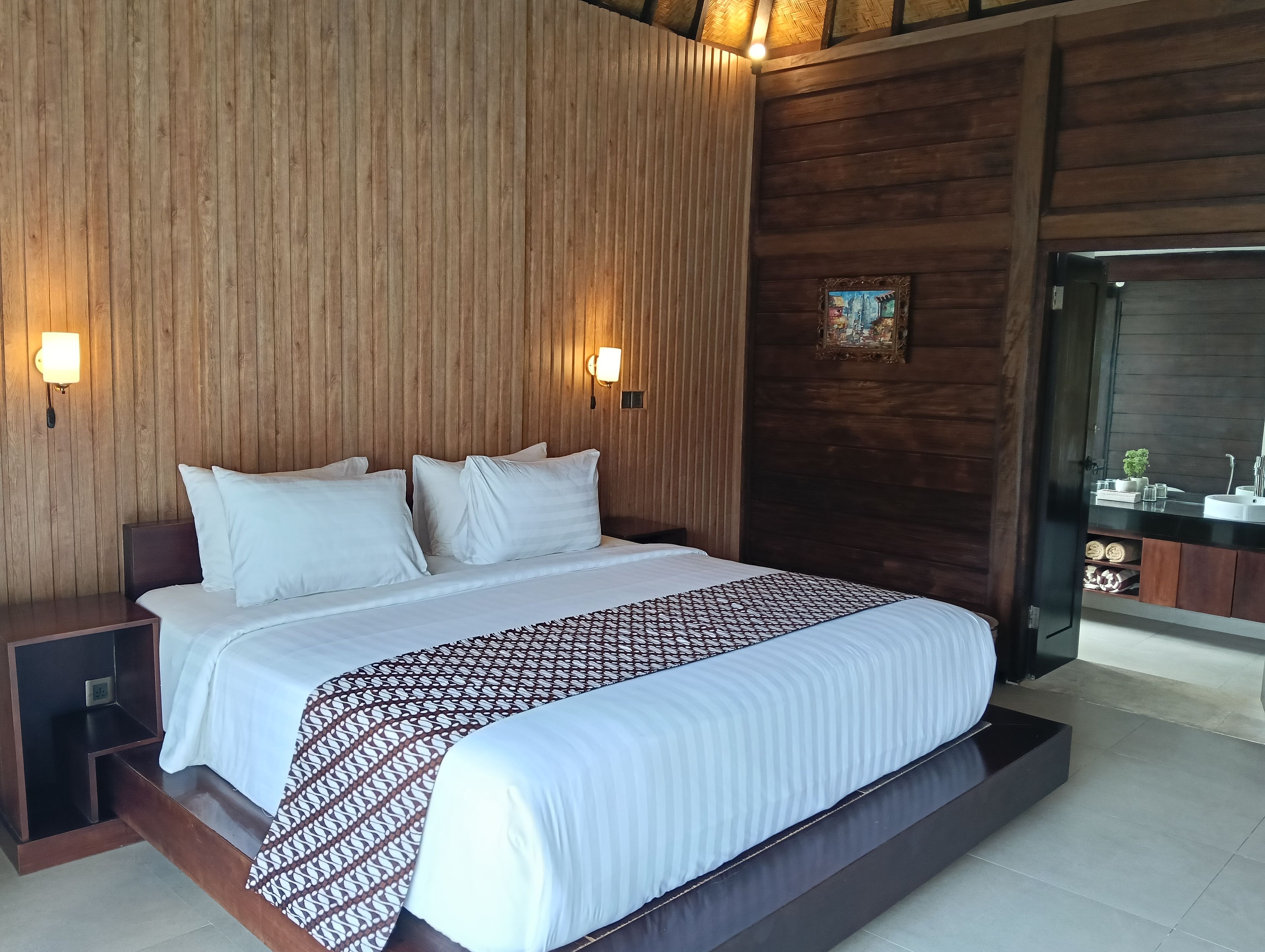 A hotel room with a large bed, wooden panel walls, two wall-mounted lights, a painting, and a bathroom visible through a glass door.