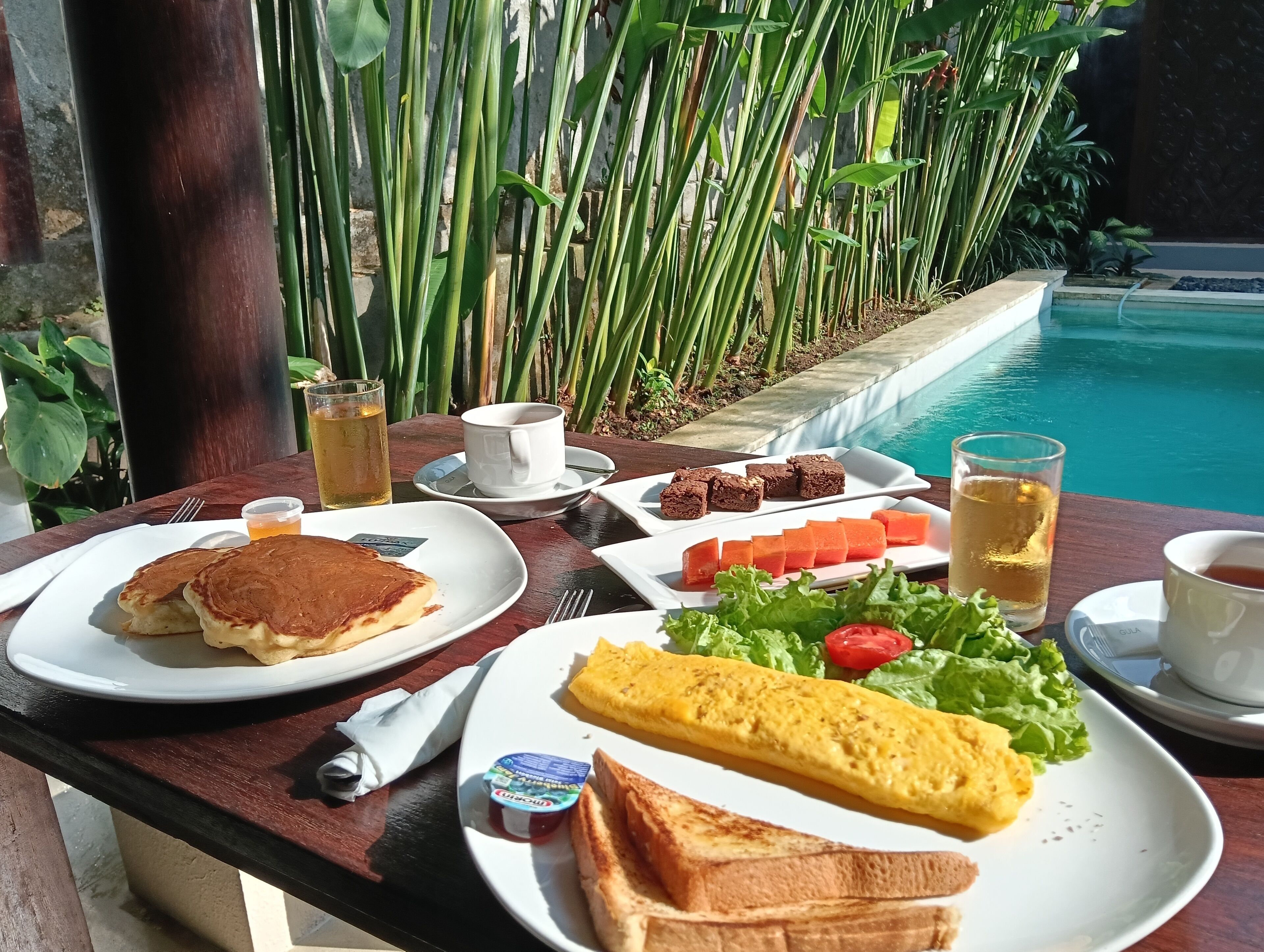 A dining table with breakfast items including pancakes, toast, omelette, and a salad, set against a backdrop of a swimming pool and greenery.