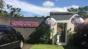 Un'auto nera parcheggiata di fronte a un edificio con un cartello che riporta la scritta 'Sri Umi Ratu Villa'. Sulla destra dell'ingresso sono presenti due ombrelli.