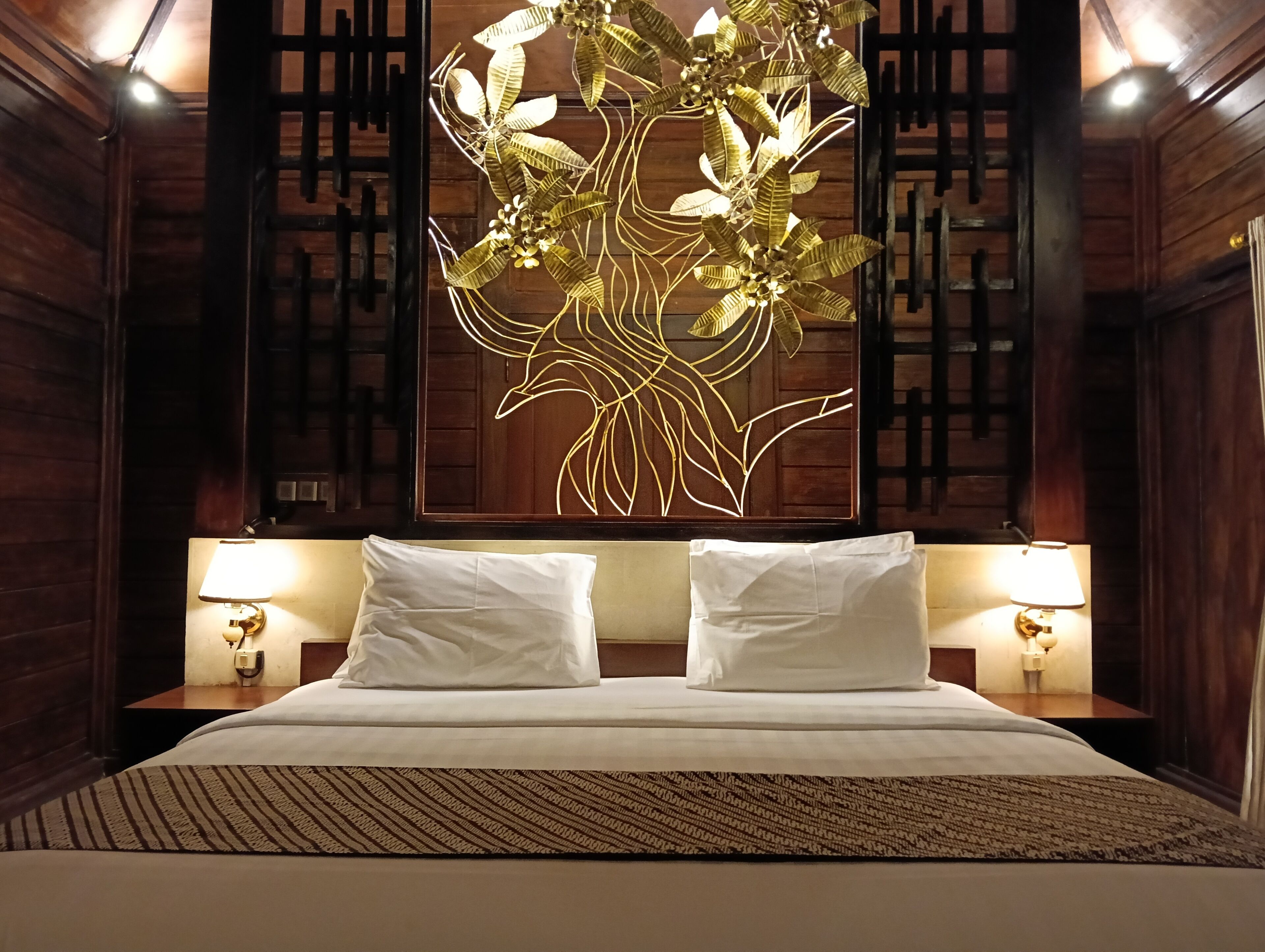 A hotel room with a bed, two bedside lamps, a wall-mounted decorative piece, and wooden paneling.