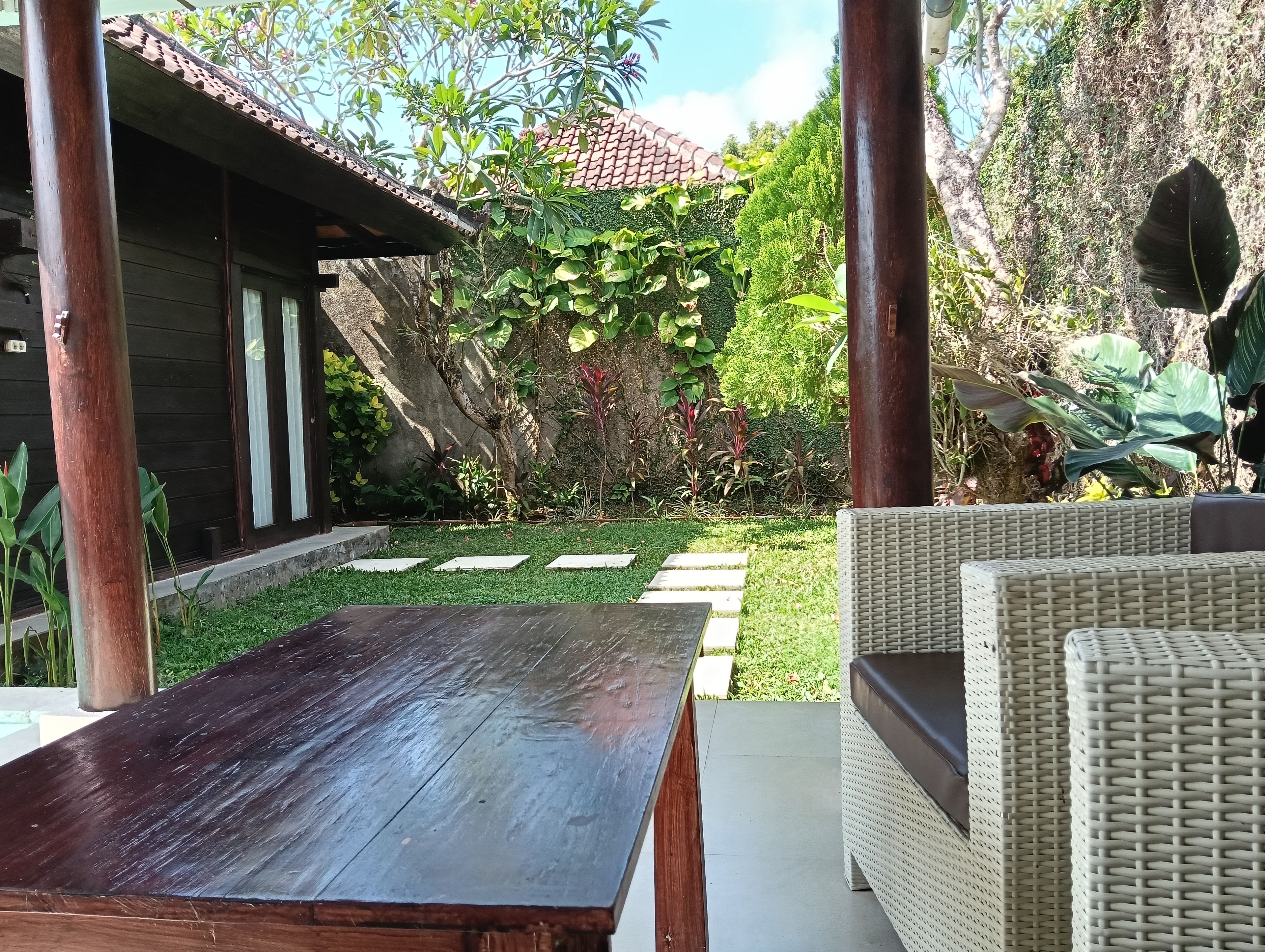 Een terras met een houten tafel en rieten meubilair, omgeven door weelderige begroeiing en een dak van tegels.
