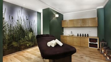 Un cuarto de spa con piso de madera, una camilla de masaje, un mural en la pared, un mueble de madera con botellas y una pequeña repisa con velas.