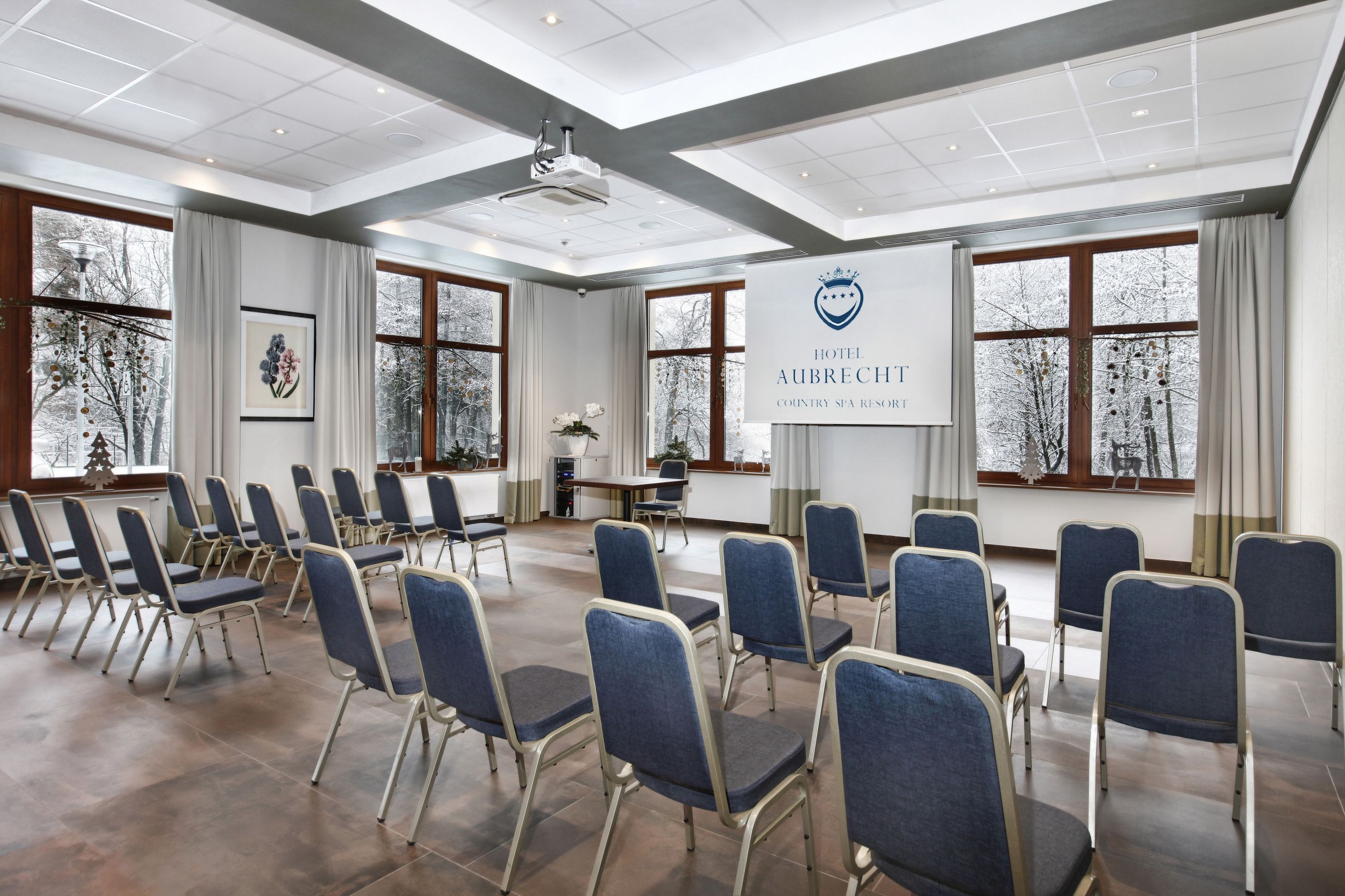 Un salón de conferencias con filas de sillas azules, un gran cartel con el nombre del hotel y amplias ventanas con vistas a árboles.