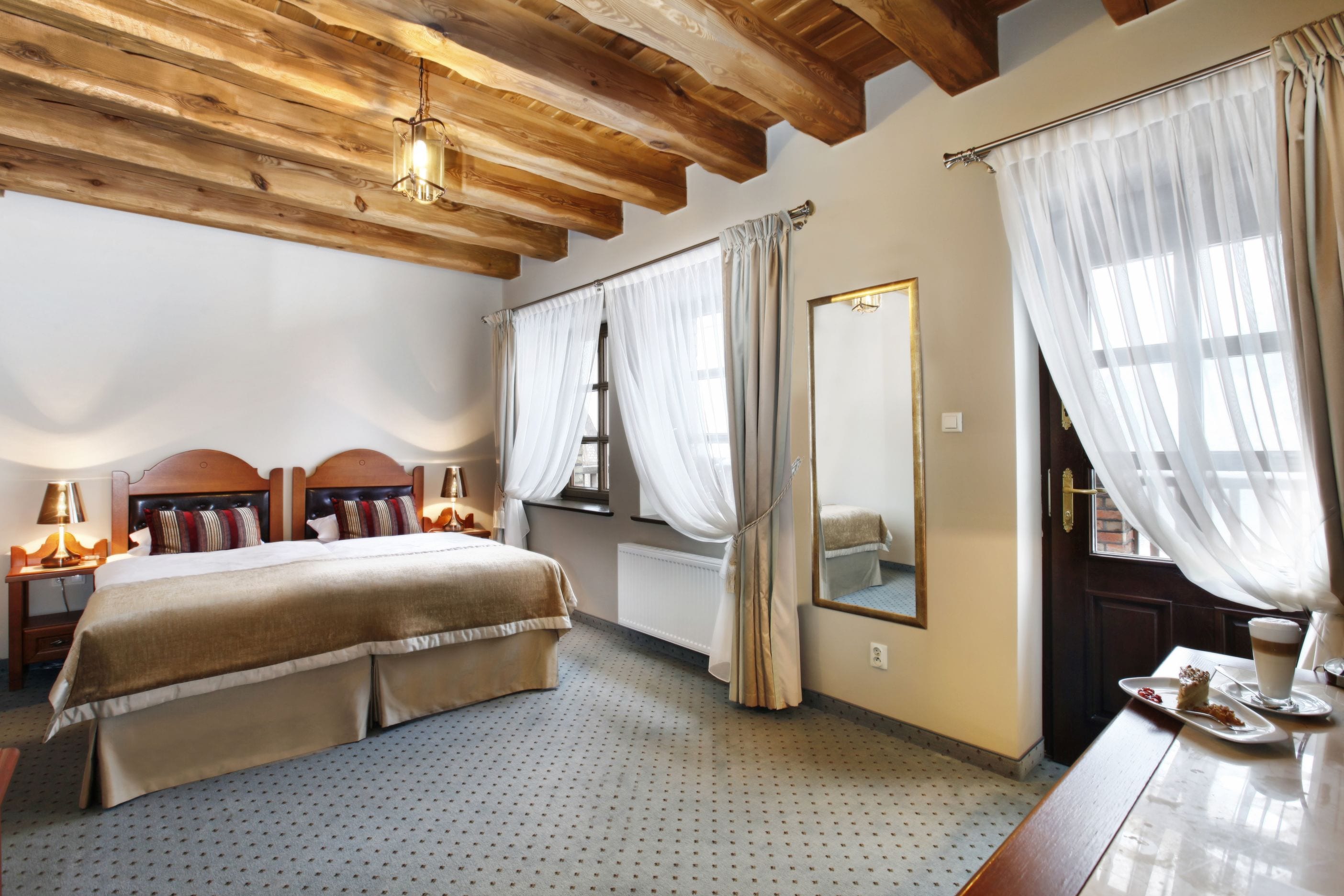 A bedroom with a wooden ceiling, a large bed with a headboard, a bedside table with a lamp, a mirror, and a window with curtains.