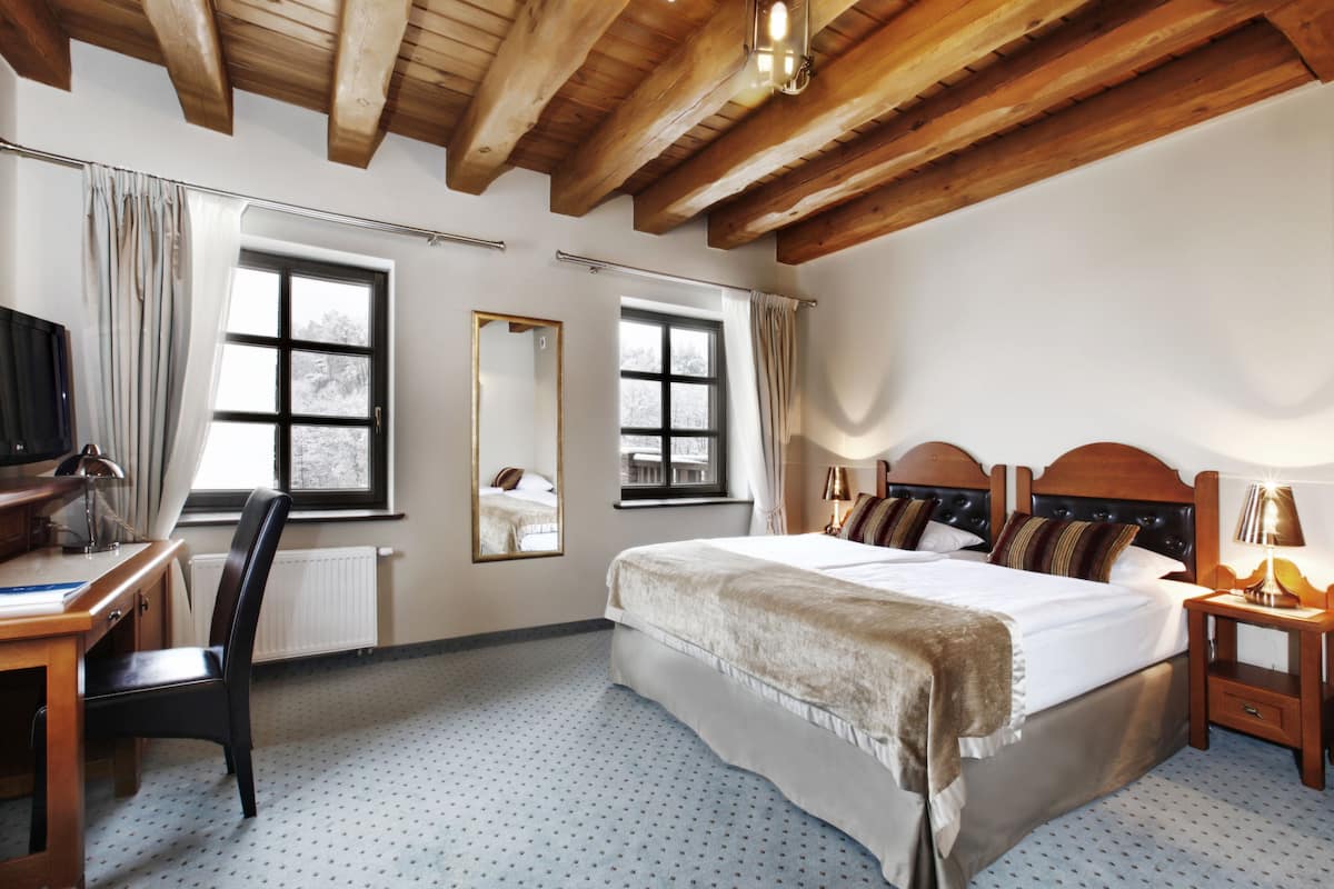 Un dormitorio con techo de madera, una cama grande con cabecera, un escritorio con silla y dos ventanas con cortinas.