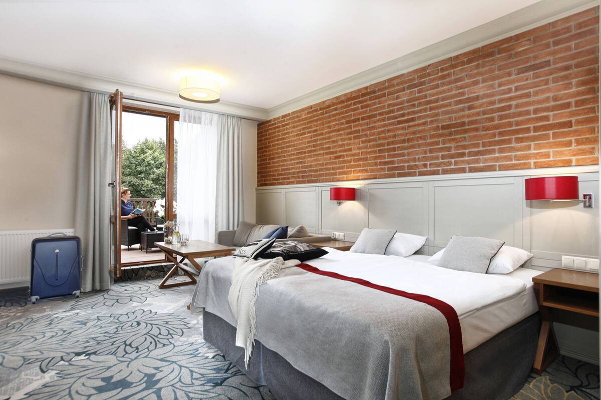 Habitación de hotel con una cama grande, un escritorio, una silla, una maleta y vistas al exterior a través de una puerta corredera.