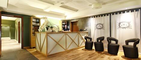 A reception area with a wooden reception desk, three black chairs, a potted plant, and a wall with framed pictures.